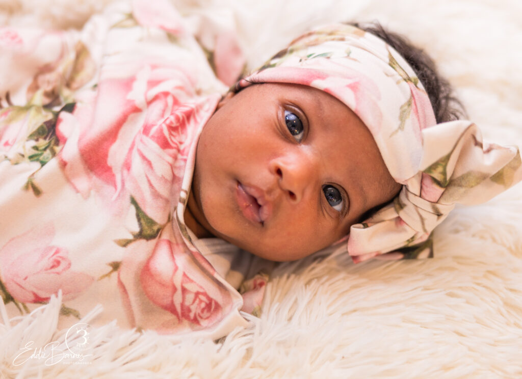 Newborn baby girl in floral outfit and matching headband lying on soft white blanket