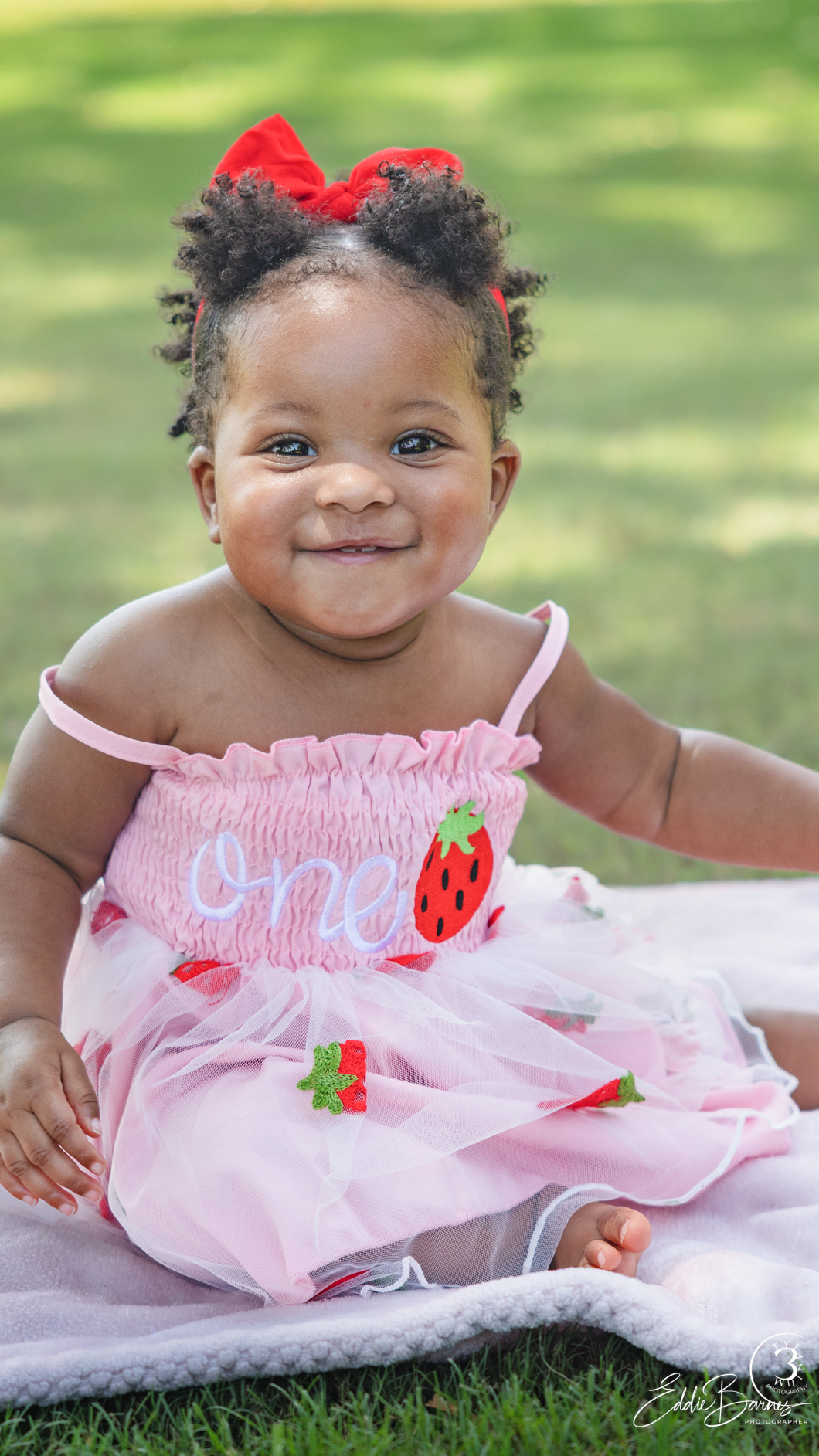Smiling baby girl in pink strawberry birthday dress with red bow sitting on blanket outdoors